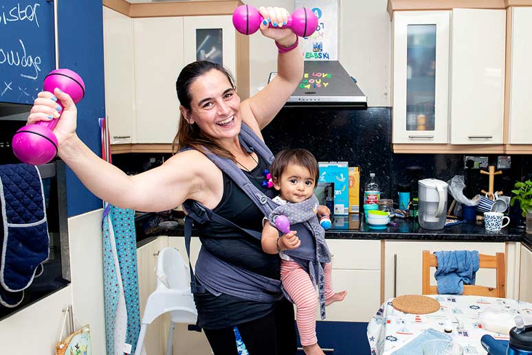 Alice doing a home workout in her kitchen with her baby daughter in a sling.